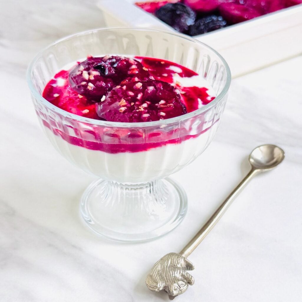 front image of bowl of yogurt topped with baked plums placed on marble along with a spoon.