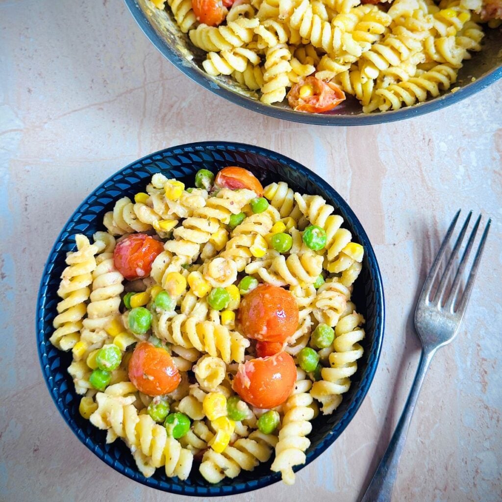 black bowl of hummus pasta with corn, peas and cherry tomatoes placed on granite along with a fork next to it and a pan of pasta behind it.