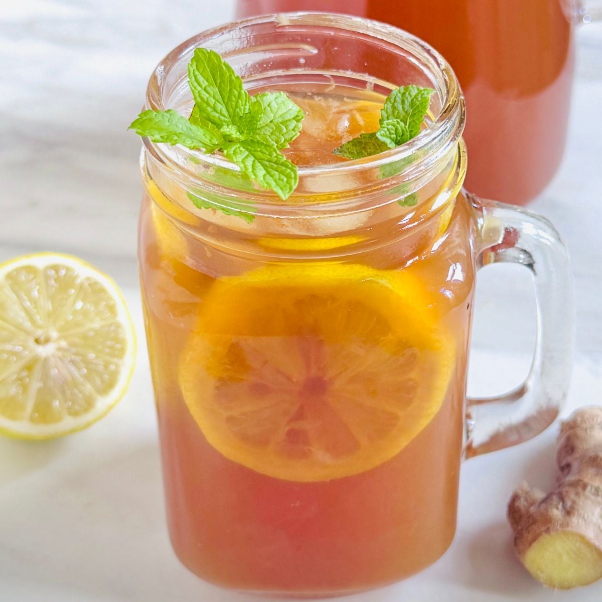 close up image of glass of spiced lemon ginger tea garnished with mint placed on marble along with a jug of tea, half lemon and ginger.