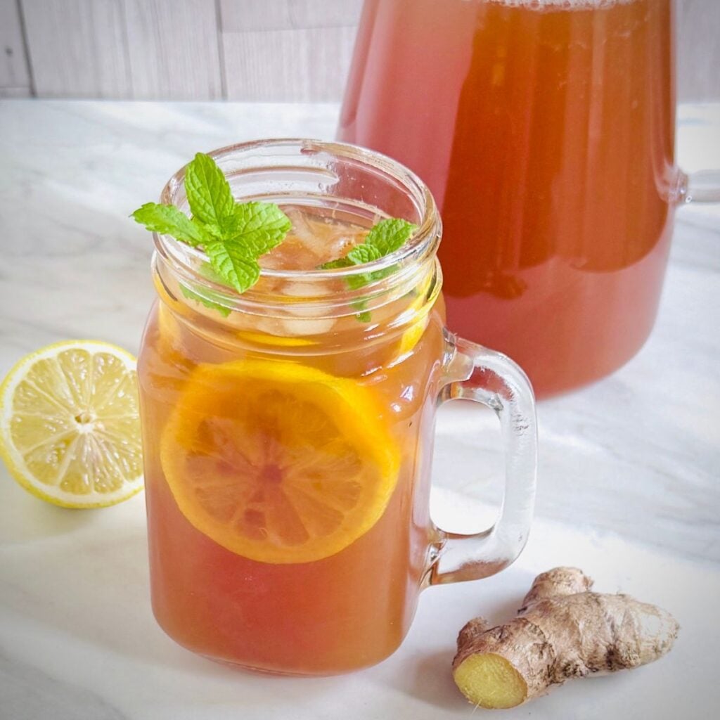 glass filled with spiced lemon ginger iced tea placed on marble along with a jug of tea, half lemon and ginger.