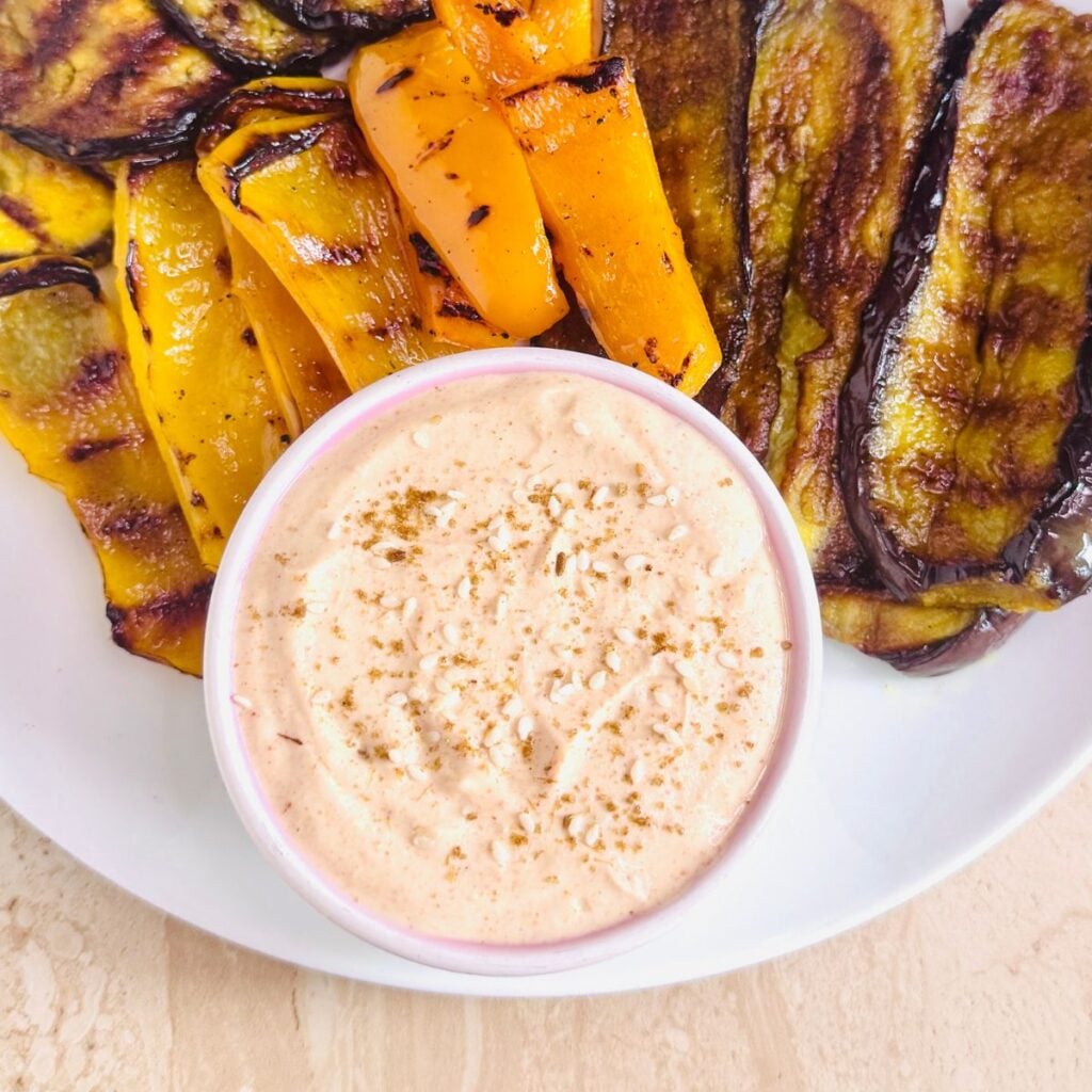white bowl of spicy tahini yogurt sauce on a plate with grilled vegetables.