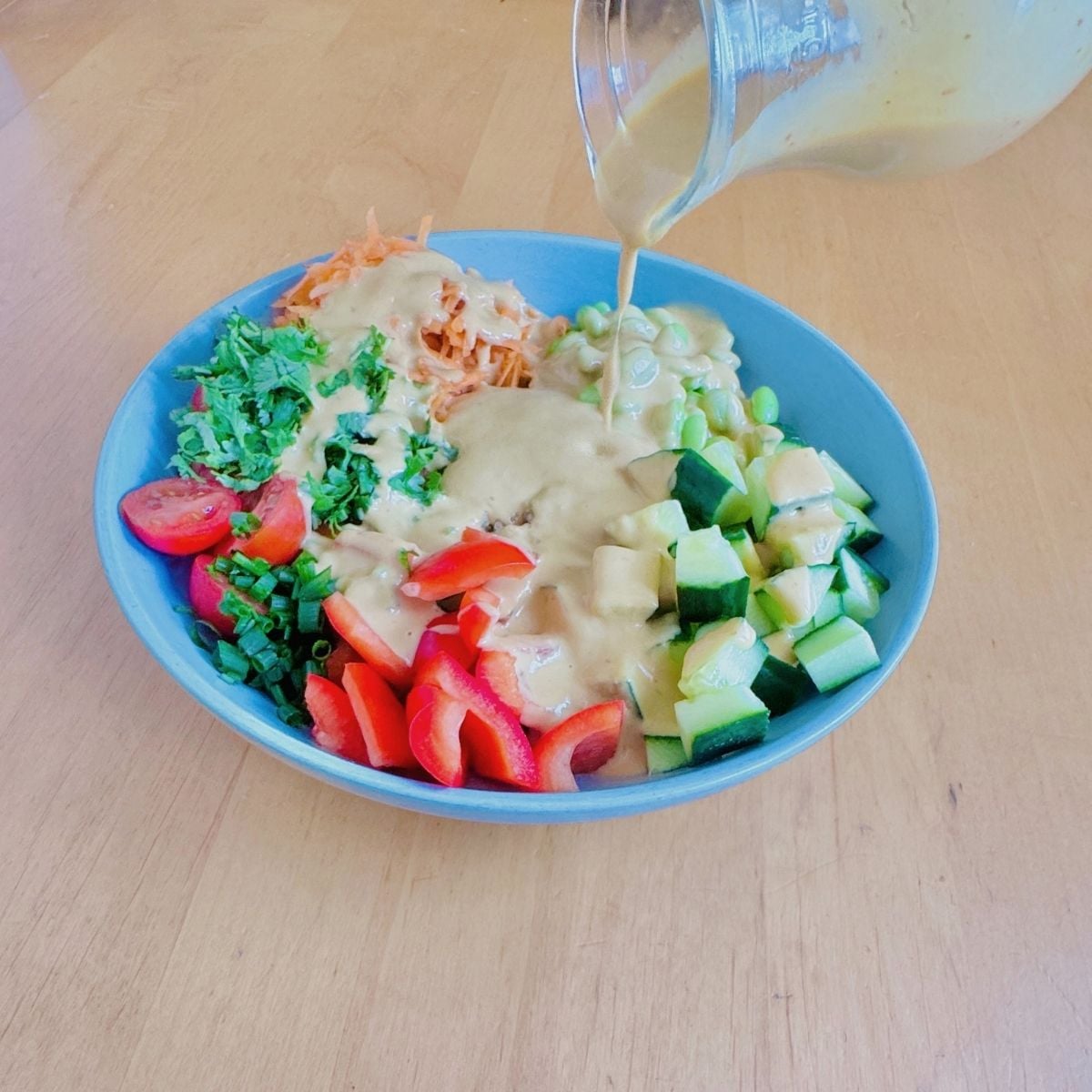 pouring mango chilli peanut butter dressing over quinoa edamame salad in blue bowl.