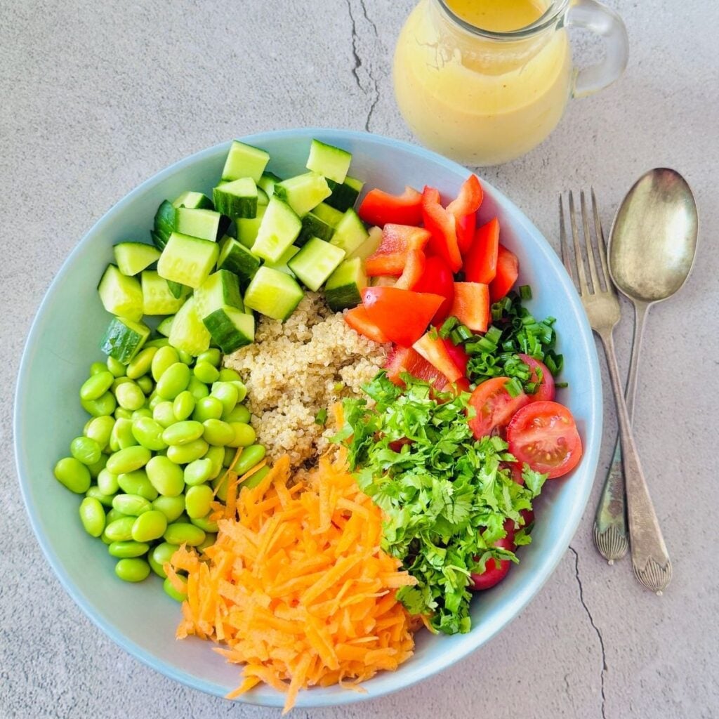 blue bowl of quinoa, edamame, vegetables placed on blue granite along with cutlery and glass jug of mango peanut butter dressing.