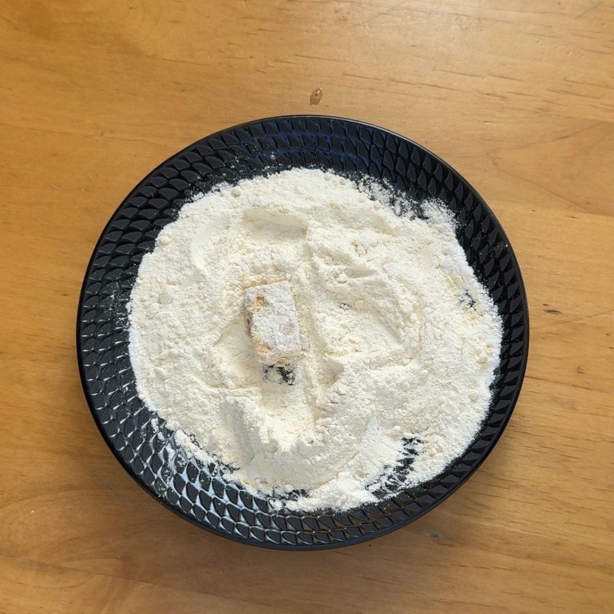 battered tofu cube on a black plate of white flour placed on wooden table