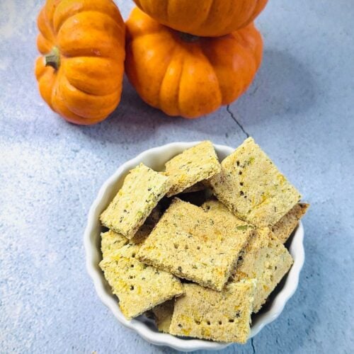white bowl of savoury pumpkin almond flour crackers placed on blue granite along with pumpkins.