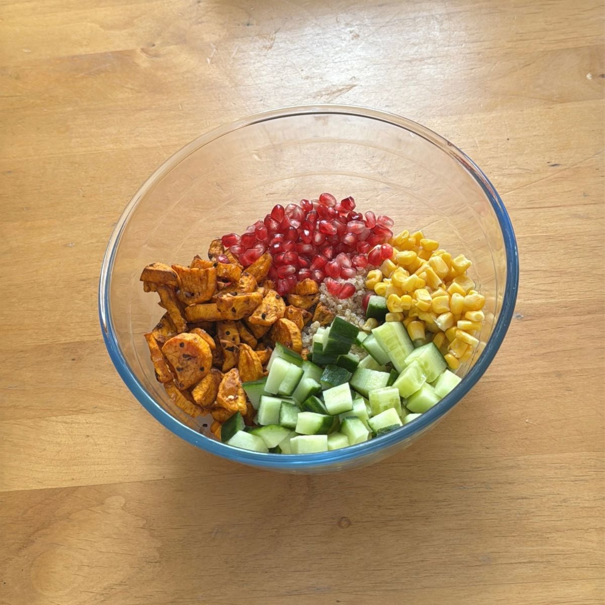 glass bowl filled with roasted sweet potato, cucumber, pomegranate, sweetcorn and quinoa placed on wooden table.